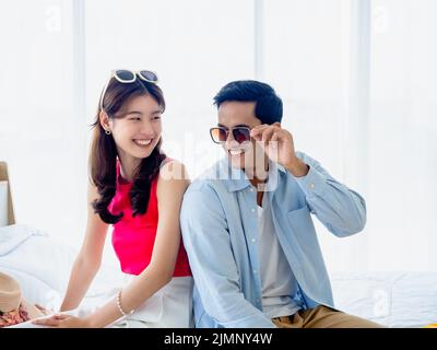 Glückliches asiatisches Paar Liebhaber im Sommer, junge Mann und Frau tragen Sonnenbrille mit Spaß Lächeln, sitzen auf dem Bett im hellen Schlafzimmer in der Nähe der Glas gewinnen Stockfoto