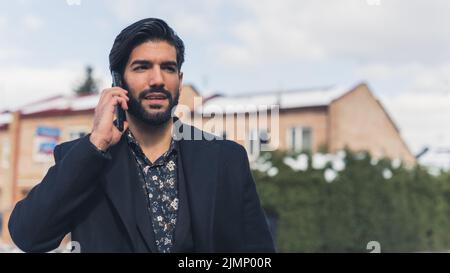 Hispanischer Mann mit glattem Haar und Bart, der Hemd und Mantel trägt, der durch die Stadt läuft und telefoniert. Geschäftsanruf. Hochwertige Fotos Stockfoto