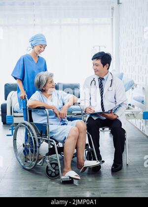 Ältere Frauen Patienten im Rollstuhl lächeln mit männlichen Chirurgen Ärzte und professionelle weibliche Krankenpflegeassistenten in weiß erholt Zimmer im Krankenhaus, V Stockfoto