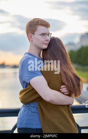 Mädchen mit langen dicken dunklen hören umarmt Rotschopf Junge in das blaue T-Shirt auf Brücke, Teenager Liebe bei Sonnenuntergang Stockfoto