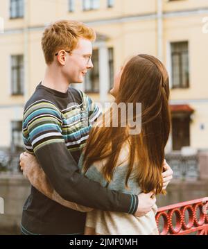 Mädchen mit langen dicken dunklen hören umarmt Rotschopf Junge in das blaue T-Shirt auf einer Brücke, junges Paar. Konzept der Teenager-Liebe und Stockfoto