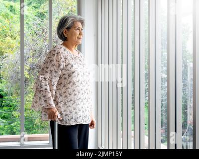 Asiatische ältere Frau, weißes Haar, das mit einem Stock steht und aus dem Glasfenster schaut, innen mit Kopierraum. Ältere Patientin mit Gehstock Stockfoto