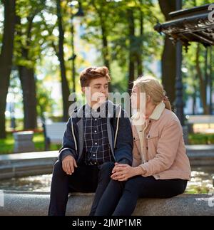 Liebhaber Junge und Mädchen schauen einander an, sitzen im Park in der Nähe des Brunnens im Herbst. Freundin und Freund reden Stockfoto
