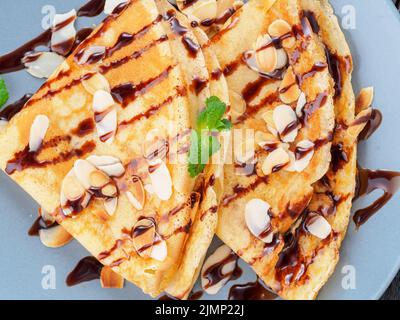 Zwei Pfannkuchen mit Schokoladensirup, Mandelflocken auf Platte, Honig fließt aus Löffel, Draufsicht, Makro Stockfoto