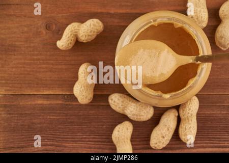 Glas Erdnussbutter und Erdnüsse in einer Schale auf braunem Holztisch, Seitenansicht, frisch zerkleinerte Erdnüsse, Draufsicht, Kopierraum Stockfoto