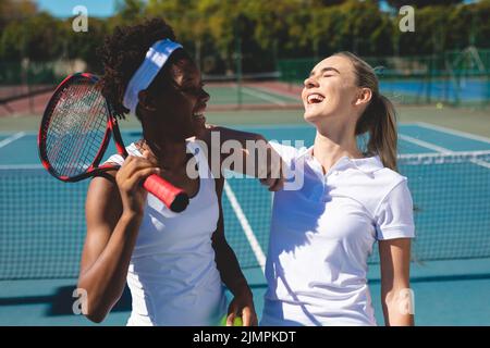 Fröhliche junge, multirassische weibliche Spieler, die am sonnigen Tag auf dem Tennisplatz stehen Stockfoto