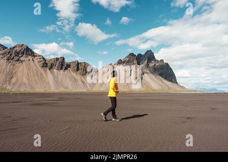 Junge asiatische Frau in gelber Jacke, die am Strand mit dem Berg Vestrahorn im wikingerdorf auf der Halbinsel Stokknes im Südosten Islands steht Stockfoto