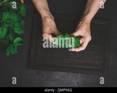 Grüne frische Minzblätter auf einem Holzbrett und zwei weibliche Hände auf einem schwarzen Holztisch, Draufsicht Stockfoto