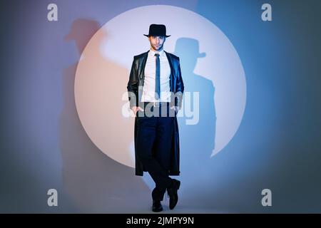 In voller Länge Porträt eines hübschen Mannes in schwarzem Mantel und Hut posiert im Studio im Rampenlicht Stockfoto