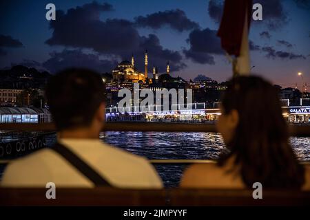 Istanbul, Türkei. 06. August 2022. Menschen, die während des Sonnenuntergangs in Istanbul auf dem offenen Gelände der Fähre der City Lines sitzen, und die Galata-Brücke mit der Suleymaniye-Moschee im Hintergrund. Kredit: SOPA Images Limited/Alamy Live Nachrichten Stockfoto