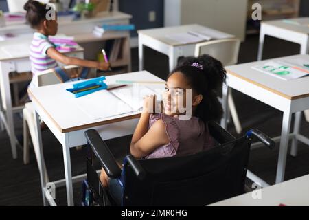 Porträt eines glücklichen biracial Grundschülerin sitzt auf Rollstuhl am Schreibtisch im Klassenzimmer Stockfoto