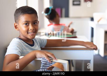 Porträt eines lächelnden, biracial Grundschüler, der im Klassenzimmer am Schreibtisch sitzt Stockfoto