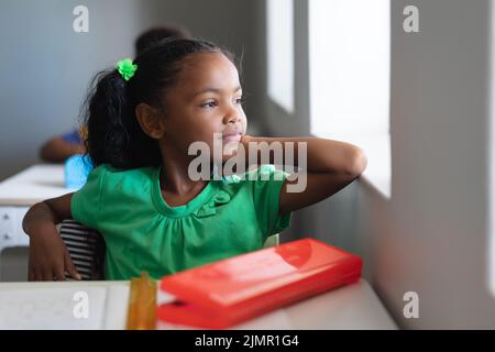 Nachdenkliches afroamerikanisches Grundschulmädchen, das durch das Fenster schaut, während es im Unterricht sitzt Stockfoto