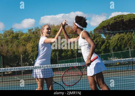 Glückliche junge, multirassische Tennisspielerinnen, die am sonnigen Tag High-Five über das Netz auf dem Platz geben Stockfoto