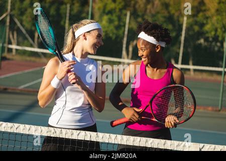 Glückliche, multirassische junge Tennisspielerinnen sprechen, während sie Schläger am Platz halten Stockfoto