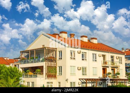 Wohnhaus mit gefliestem Dach und großen Terrassen mit Holzelementen, Glasbalkonen und dekorativen, belaubten, blühenden Pflanzen, Liegestühlen und Cha Stockfoto