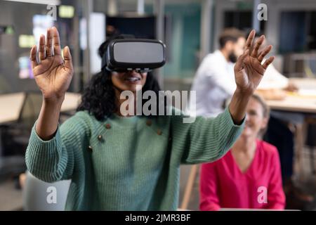 Lächelnde junge afroamerikanische Geschäftsfrau trägt Virtual Reality-Simulator gestikuliert im Büro Stockfoto