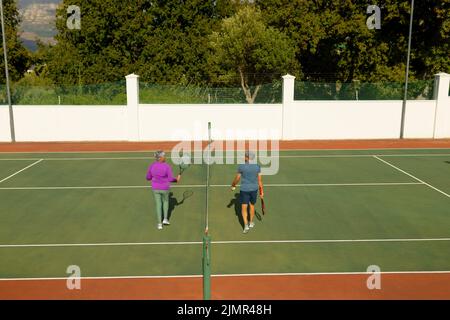 Rückansicht des älteren Birazialmannes mit einer älteren Frau, die während eines sonnigen Tages auf dem Tennisplatz im Netz herumläuft Stockfoto