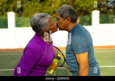 Seitenansicht eines liebenden Senioren-Paares mit geschlossenen Augen, die sich küssen, während sie auf dem Tennisplatz stehen Stockfoto