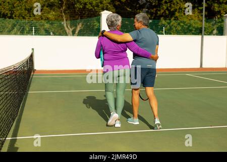 Rückansicht des älteren Biracial-Mannes und seiner Frau mit den Armen, die am sonnigen Tag auf dem Tennisplatz herumlaufen Stockfoto