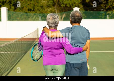 Rückansicht der älteren Frau und des Mannes mit den Armen, die an sonnigen Tagen auf dem Tennisplatz standen Stockfoto