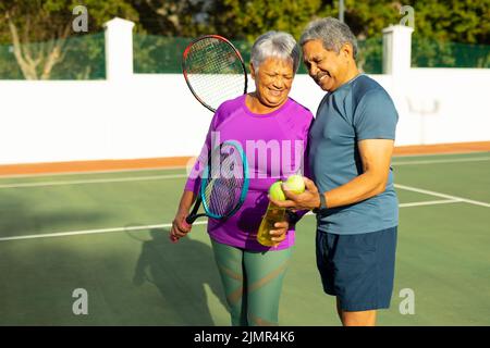 Glückliches Senioren-Paar mit Tennisbällen, Schlägern und Wasserflasche auf dem Tennisplatz Stockfoto