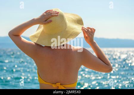 Die elegante Dame im gelben Bikini bewundert das glitzernde Meer im Resort bei Sonnenlicht. Junge Frau hält Strohhut mit den Händen an windigen Sommertagen Nahansicht Stockfoto