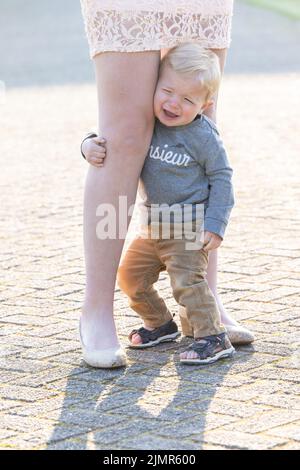 Trauriger kaukasischer blonder kleiner Junge, der ihre Mutter an den Beinen umarmt Stockfoto