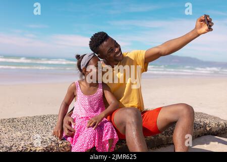 Glücklicher afroamerikanischer junger Vater, der Selfie mit dem Mädchen über das Smartphone macht, während er auf dem Felsen sitzt Stockfoto