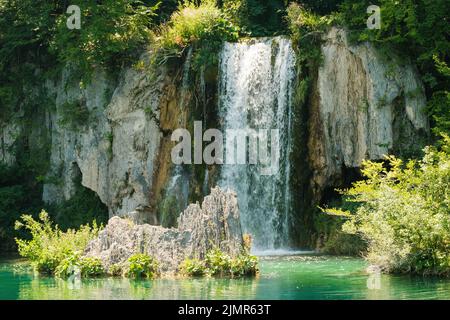 Der Wasserfall fließt bei Sonnenlicht von einer felsigen Klippe in klares türkisfarbenes Wasser. Atemberaubende Aussicht auf die Aquamarin-Kaskade im Nationalpark auf den Plitvicer Seen Stockfoto