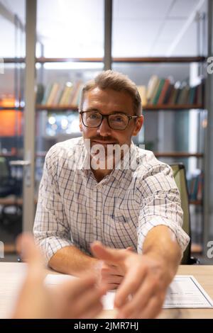 Porträt eines selbstbewussten kaukasischen Geschäftsmannes, der sich die Hände schüttelt, während er mit Zustimmung am Schreibtisch sitzt Stockfoto