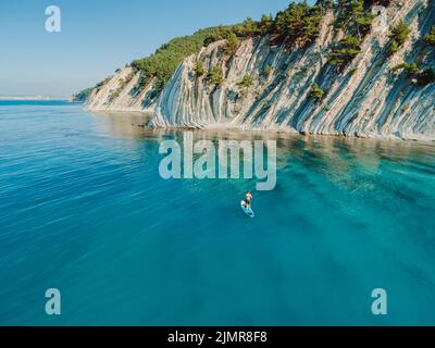 Mann auf Stand Up Paddle Board im blauen Meer. Surfer wandern auf SUP-Board in ruhigem Meer mit unglaublicher Landschaft. Luftaufnahme Stockfoto