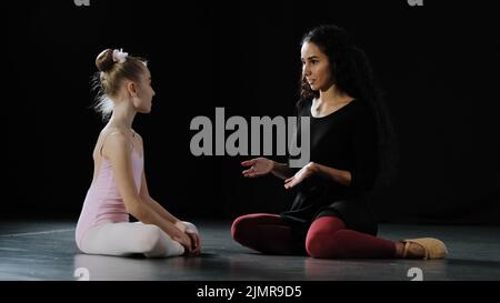 Kleines Mädchen Ballerina Tänzerin Anfänger akrobaten Kind in Trikot sitzt auf dem Boden in der Tanzklasse mit erwachsenen Frau Lehrer Trainer zuhören Beratung Gespräche Stockfoto