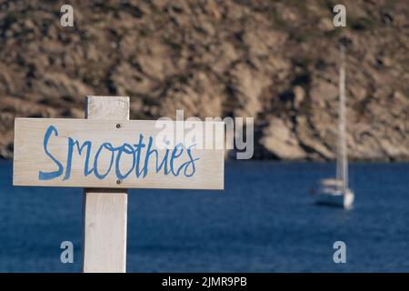 Blick auf Holzschilder für Smoothies und Live-Musik und das Meer im Hintergrund am Strand von Mylopotas in iOS Griechenland Stockfoto