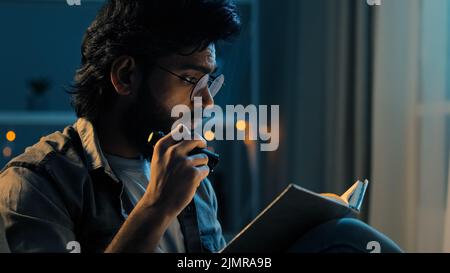 Arabisch-indischer erwachsener Mann mit Bart, der eine Brille trägt, sitzt nachts zu Hause, spät nachts, Dunkelheit, Verdunkelung, liest spirituelles Buch, Lehrbuch mit Taschenlampe hom Stockfoto