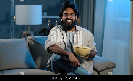Arabischer erwachsener bärtiger Mann in Brille indischer männlicher Junggeselle sitzt nach der Arbeit auf dem Sofa im dunklen Wohnzimmer schaltet den Fernseher ein, wobei die Fernbedienung den Sender auswählt Stockfoto