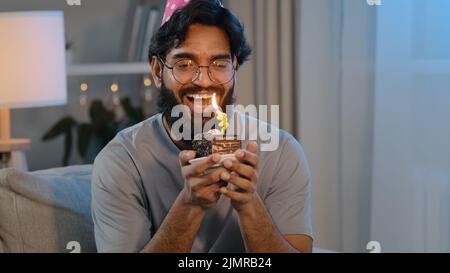 Portrait fröhlich fröhlich arabisch indisch bärtig Mann in Gläsern in festlicher Mütze zu Hause Abend feiert Jubiläum Spaß mit kleinen Kuchen halten Stockfoto