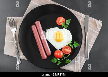 Herzförmige Rühreier mit Würstchen auf schwarzem Teller. Stockfoto