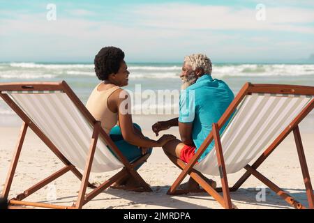 afroamerikanisches Paar, das sich unterhielt, während es auf Liegestühlen am Sandstrand gegen den bewölkten Himmel saß Stockfoto