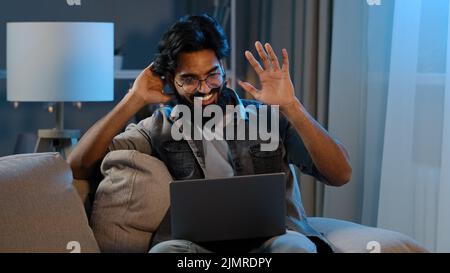 Arabisch indisch bärtig Mann Millennial guy Geschäftsmann Freiberufler sitzen zu Hause Sofa in der Nacht dunkel spät winkt hallo in webcam online Stockfoto