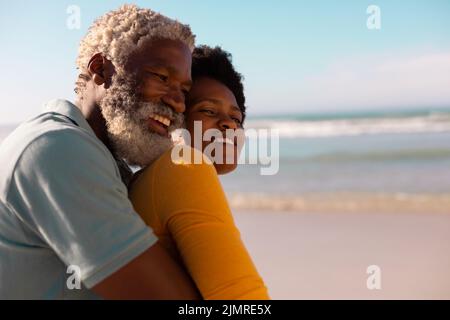 Seitenansicht eines glücklichen bärtigen afroamerikanischen Senioren, der eine reife Frau gegen Meer und Himmel umarmt Stockfoto