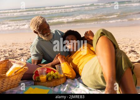 Fröhliche afroamerikanische reife Frau, die auf dem Schoß eines älteren Mannes bei Essen und Trinken auf dem Meer liegt Stockfoto