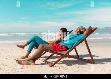 Seitenansicht eines afroamerikanischen Paares, das sich im Sommer auf Liegestühlen vor dem Meer und dem blauen Himmel entspannen kann Stockfoto