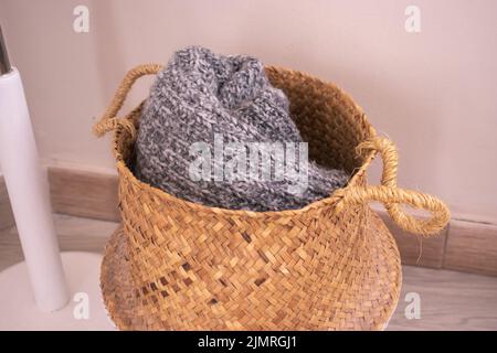 Warmer grauer gestrickter Winterschal aus Pullover in einem braunen Korbkorb auf dem Boden. Stockfoto