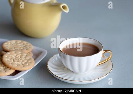 Eine frisch gebrühte Tasse Tee in einer feinen Bone-china-Tasse und Untertasse.der Tee wird mit zwei reichen Tee-Keksen serviert und die Teekannen, die für die Herstellung verwendet werden, sind im Blickfeld Stockfoto