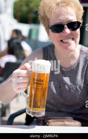 Eine Kundin, die an einem Tisch vor einer Bar sitzt. Sie sitzt bei strahlendem Sonnenschein und hält ein volles Glas Estrella Galicia Lager in voller Pint-Größe. Stockfoto