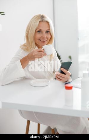 Kaukasische Senior schöne Frau Client trinken Kaffee und mit Smartphone in der Beauty-Klinik Stockfoto