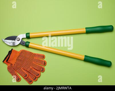 Großer Gartenscherer zum Beschneiden von Ästen an Bäumen und orangefarbene Textilhandschuhe auf grünem Papierhintergrund, Draufsicht Stockfoto