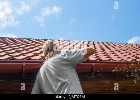 Ein Mann reinigt die Rinne eines Abflussrohres von trockenen Blättern und Trümmern. Vermeidung von Stauungen und Bruch des Abflusses der Rinne. Stockfoto