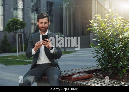 Lächelnde Büroangestellte ruhen sich während der Pause außerhalb des Büros aus und blättern in den sozialen Medien am Telefon Stockfoto